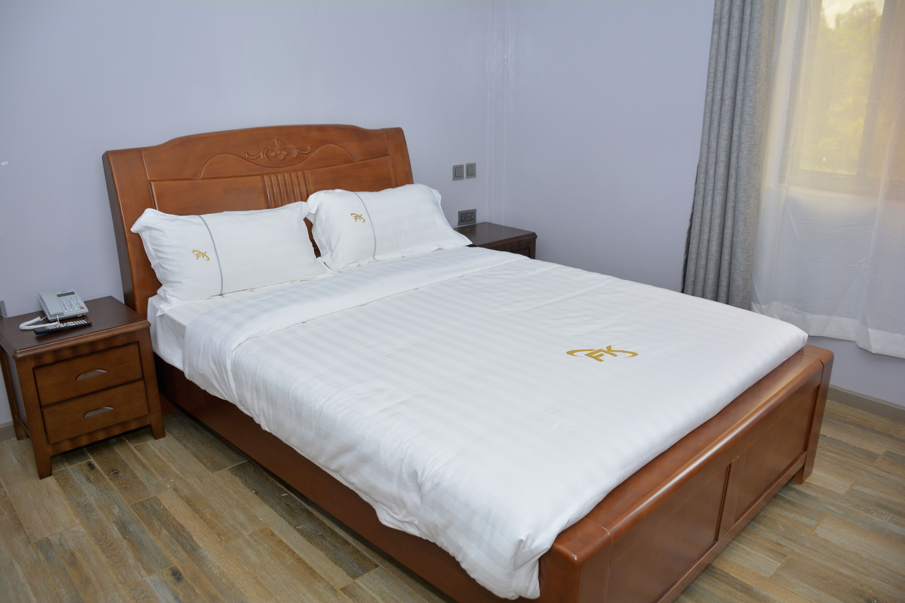 A neatly made bed with a wooden headboard, a bedside table with a phone, and a window with curtains.