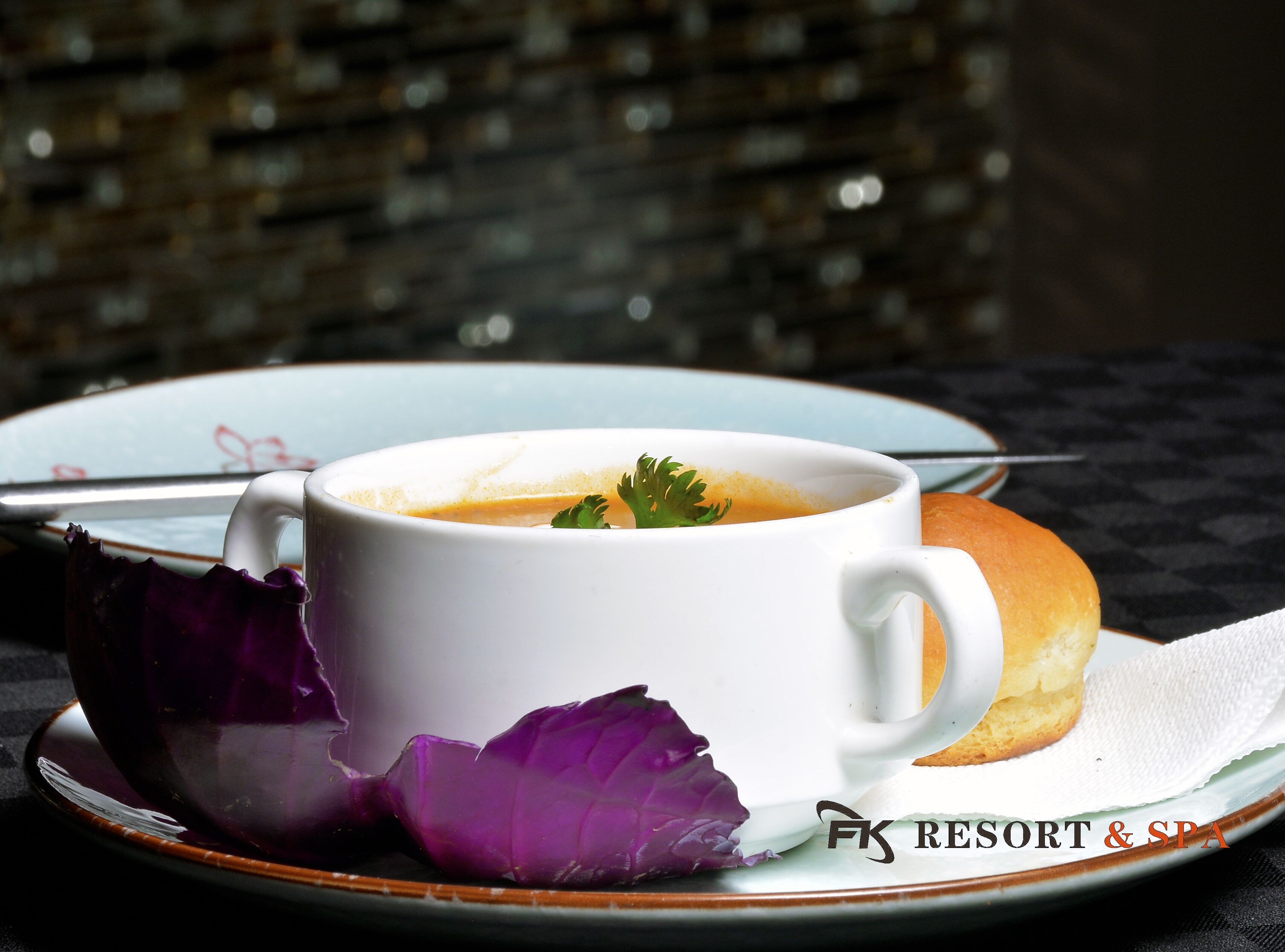A white cup of soup with a garnish on a plate, accompanied by a roll and purple leaves, on a table with a patterned tablecloth.