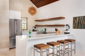 A modern kitchen with a stainless steel refrigerator, white countertops, wooden stools, and a hanging light fixture. - Villa Beach Shack (Providenciales)