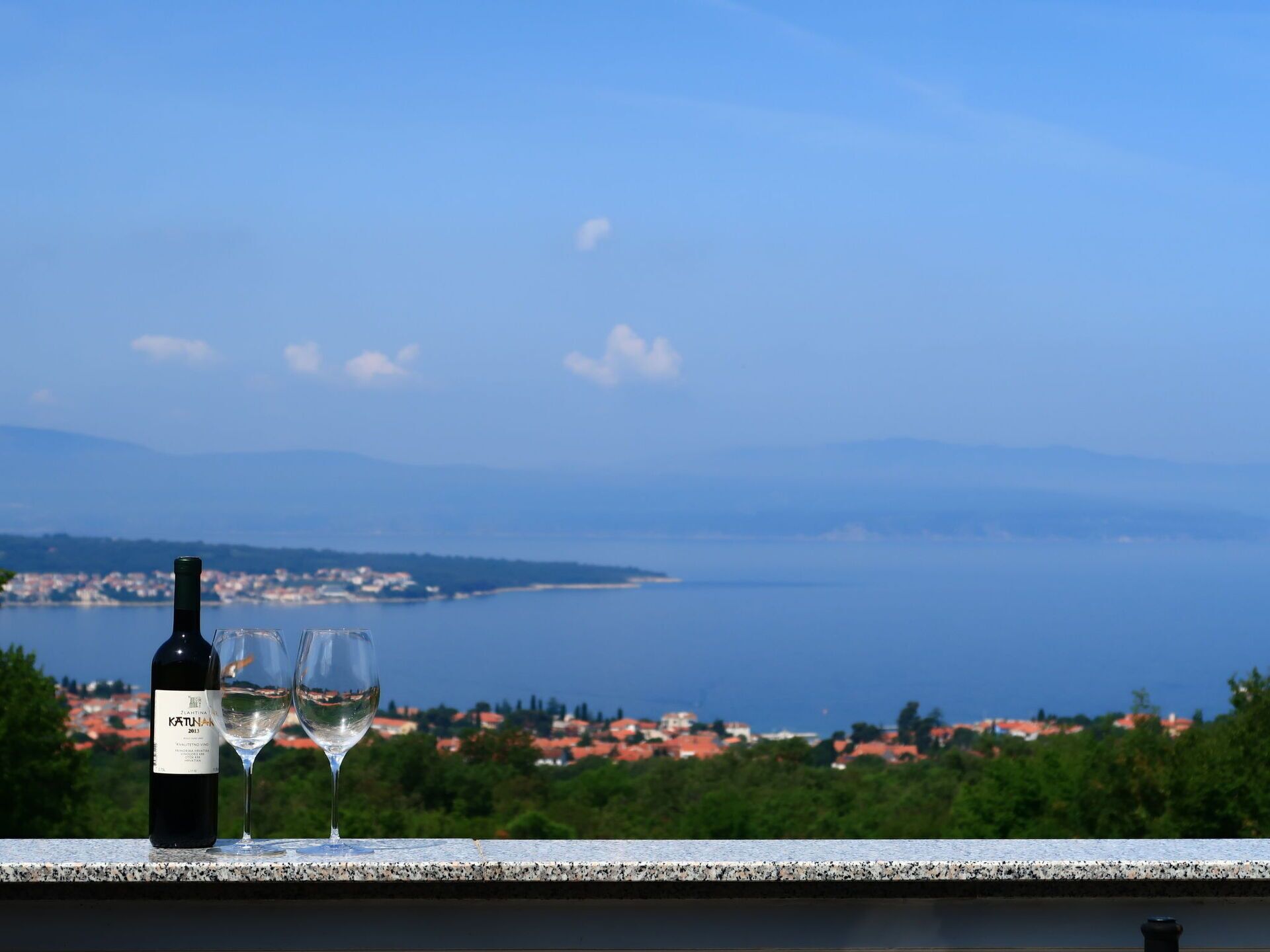 A bottle of wine and two glasses on a balcony overlooking a coastal town.