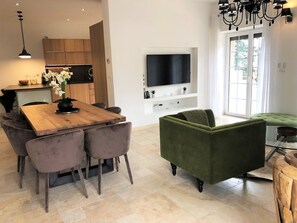 Una cocina moderna con armarios de madera, una mesa de comedor con sillas, un televisor de pantalla plana montado en la pared y un sillón tapizado en tono verde en la sala de estar.