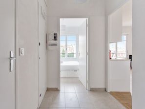A bright hallway leading to a bedroom with a window, a control panel, and a view of the city. - Appartement Libération - Welkeys (Nice)