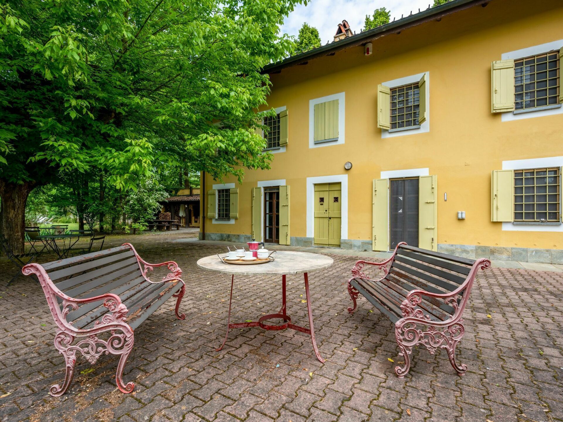 Une cour avec deux bancs décoratifs, une table ronde sur laquelle se trouve un plateau avec des tasses et des soucoupes, et un bâtiment jaune aux volets verts.