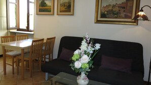 Uma sala de estar com um sofá preto, uma mesa de centro de vidro, uma mesa de jantar com cadeiras e um vaso de flores. Há quadros nas paredes e um abajur sobre o sofá.