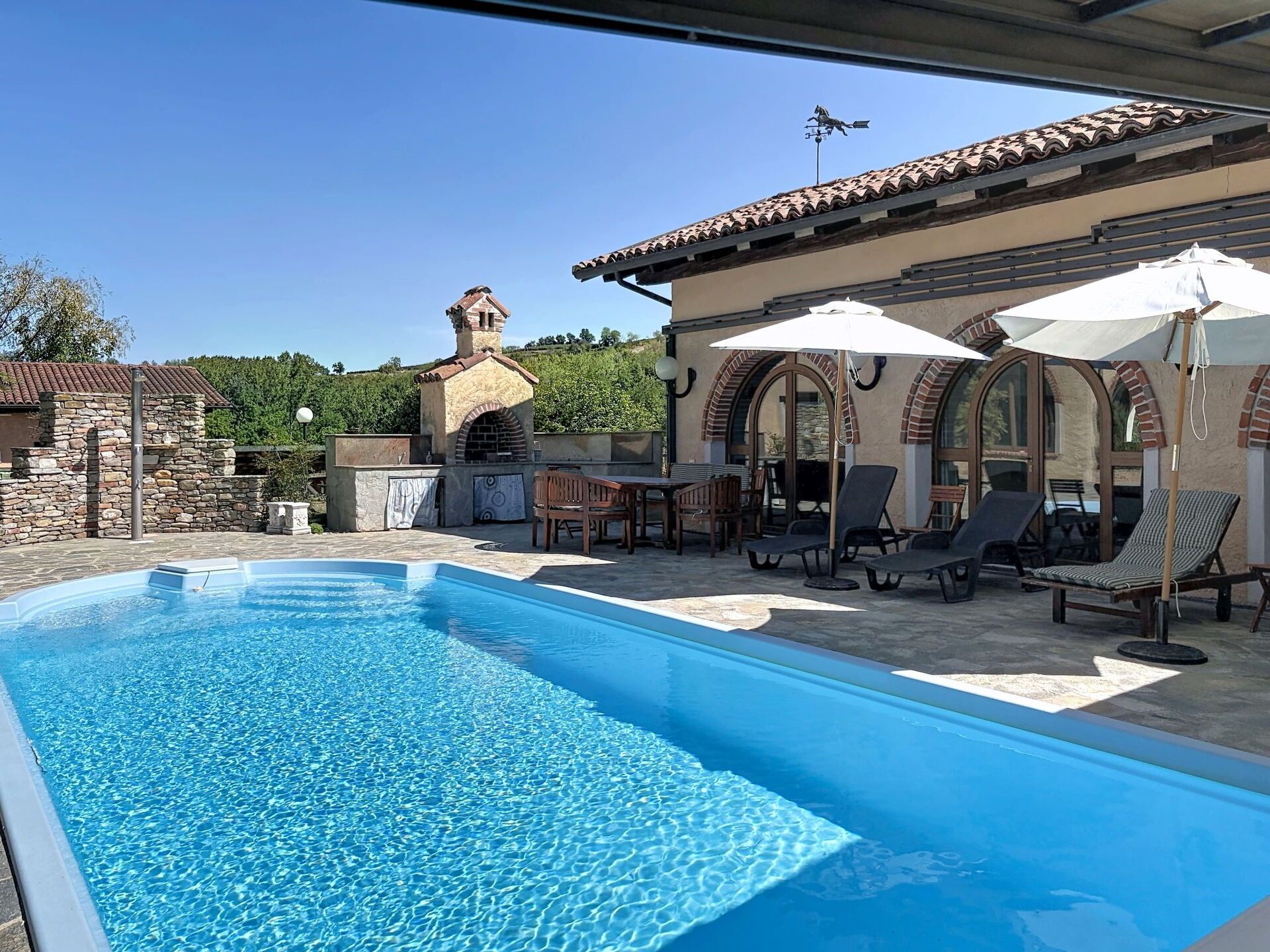 A swimming pool with lounge chairs and umbrellas, a stone building with a chimney, and a clear blue sky.