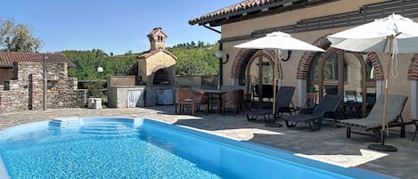 Una piscina amb gandules i ombrel·les, un edifici de pedra amb una xemeneia, i un cel blau clar.