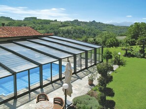 Ein von Glas umgebener Swimmingpool mit Terrassenbereich, umgeben von einem üppigen Garten und Blick auf sanfte Hügel.