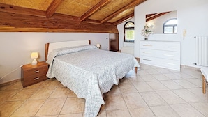 Een slaapkamer met een houten plafond, een bed met een beddekleed met dessin, een nachtkastje, een dressoir en een raam met uitzicht.