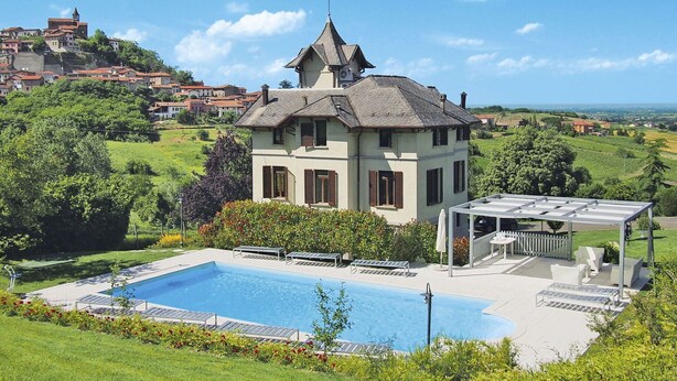 A two-story house with a swimming pool, surrounded by lush greenery and a view of a hillside village.