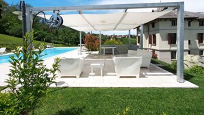 A covered outdoor seating area with a pool, white furniture, and a view of a house in the background.