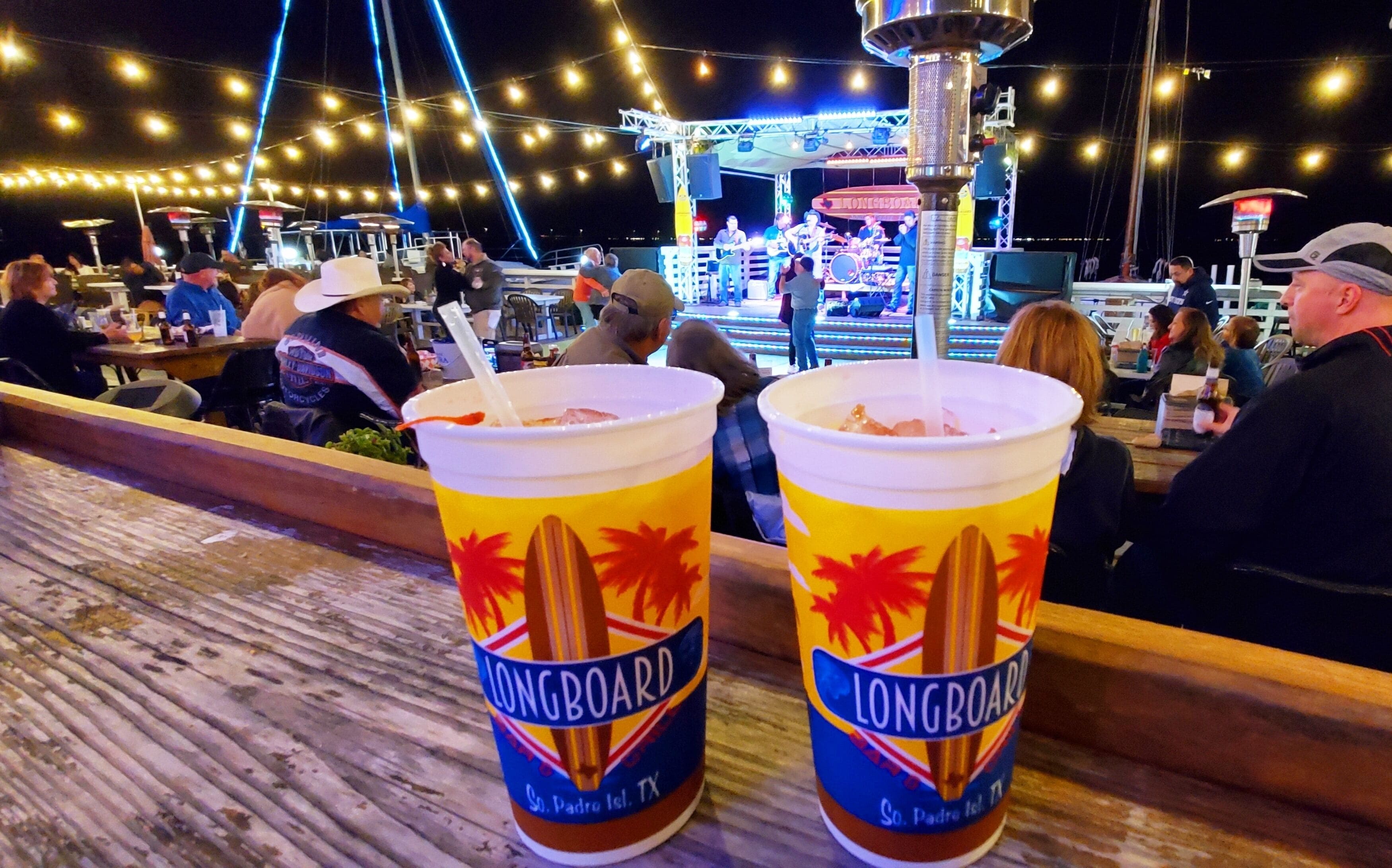 Two Longboard drinks on a wooden bar counter at a waterfront venue with a stage and audience in the background.