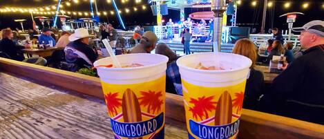 Two Longboard drinks on a wooden bar counter at a waterfront venue with a stage and audience in the background.