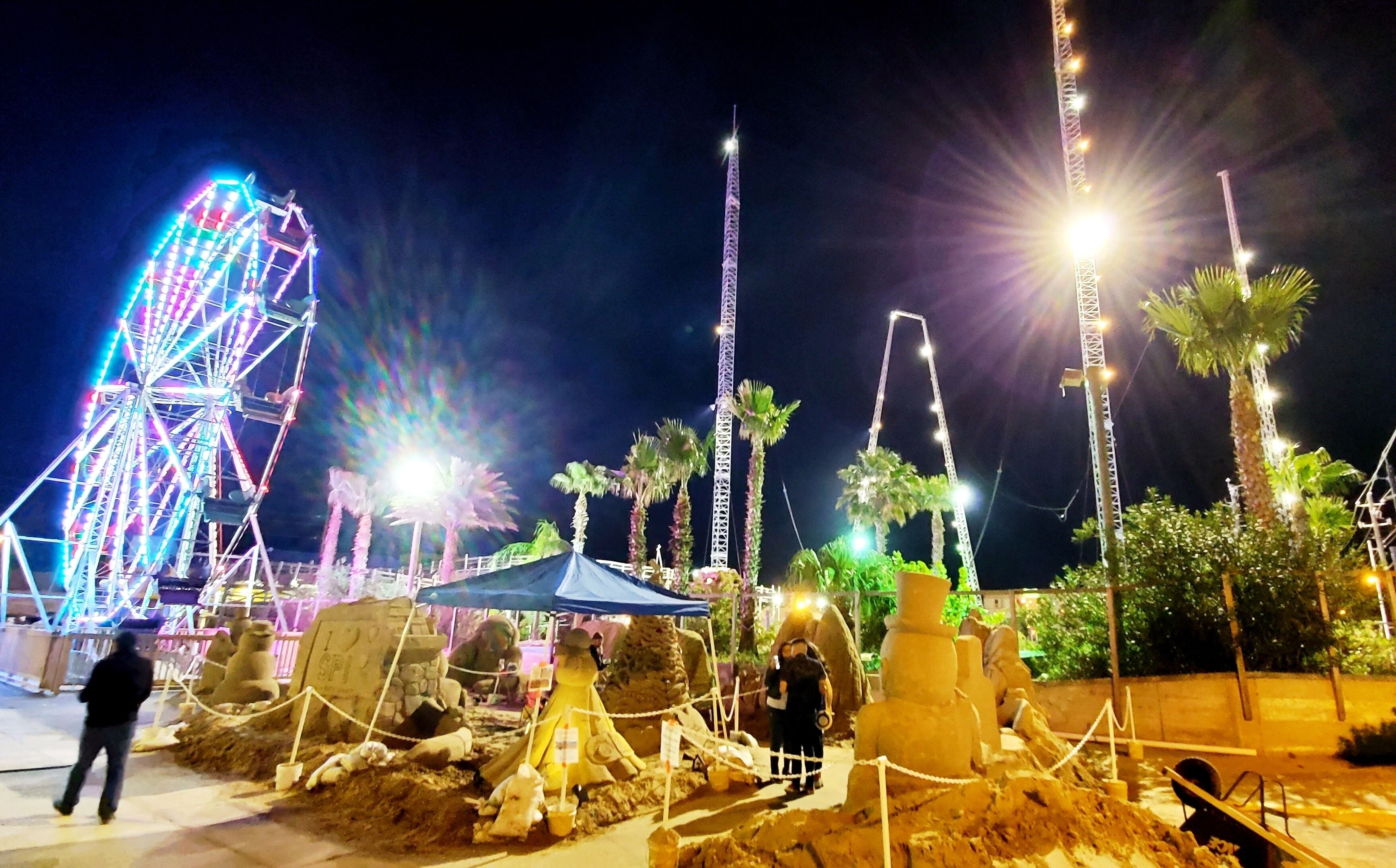 A nighttime amusement park with a Ferris wheel, palm trees, and illuminated structures.