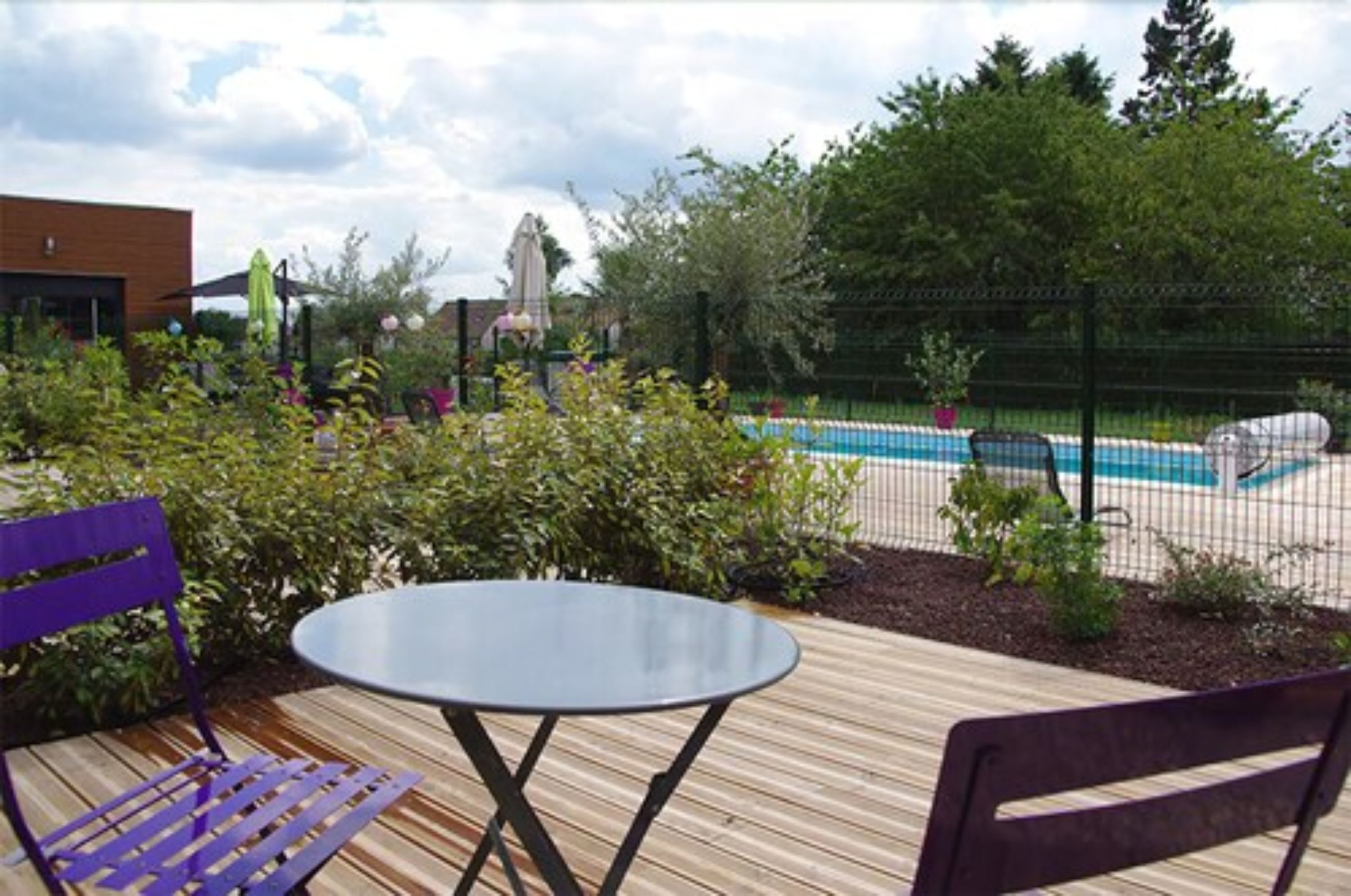 A wooden deck with a table and chairs, a swimming pool, and surrounding greenery.