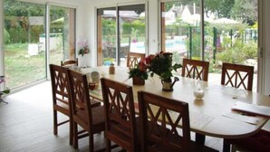 A dining area with a large table and chairs, large windows, and a view of a garden and pool. - Les Abris du Chateau (Mer)
