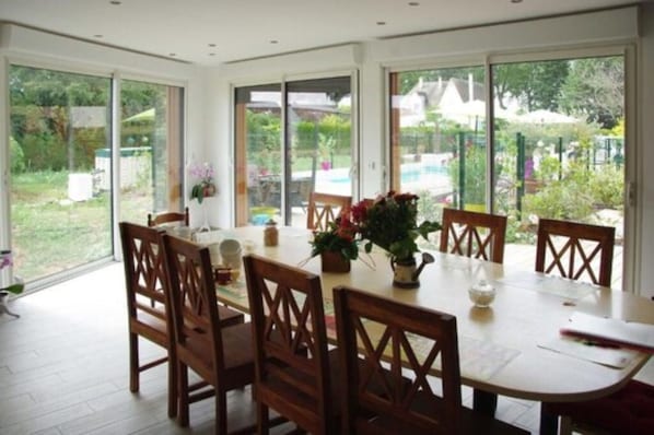 A dining area with a large table and chairs, large windows, and a view of a garden and pool.