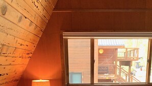 Tunnelmallinen huone, jossa on kalteva katto, valkoisilla lakanoiden ja kahden tyynyn peitetty sänky, yöpöytä lampun kanssa ja ikkuna, josta avautuu näkymä ulkona olevaan puurakennukseen.