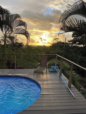 A wooden deck with a pool, a lounge chair, and a view of the sunset.