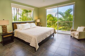 A bedroom with a bed, nightstands, a chair, and a balcony with a view of palm trees.