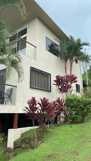 A modern two-story building with a balcony, a window with a metal grill, and a well-maintained garden with various plants.