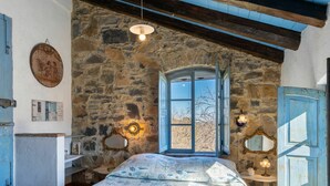 Un dormitorio con pared de piedra, una cama con colcha estampada, una ventana con vistas y una puerta azul.