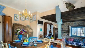 Un comedor rústico con una mesa de madera dispuesta para cuatro personas, rodeada de sillas azules. Se ven una pared de piedra y un mueble de madera al fondo.