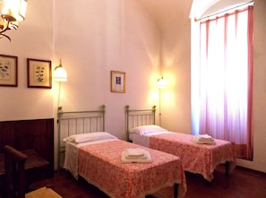 A room with two single beds, each with a floral bedspread and a white pillow. There are wall-mounted lamps, a framed picture on the wall, and a window with curtains.