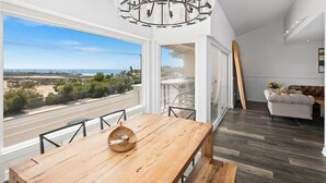 Un espace de repas moderne avec une table et des chaises en bois, une vue sur l’océan, et un salon comprenant un canapé et un tableau.