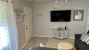 A living room with a flat-screen TV mounted on the wall, a glass coffee table, a sofa, and a patterned rug. - Located right outside of the Vibe District at the Virginia Beach Oceanfront (Virginia Beach)