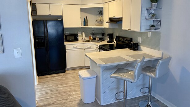 A modern kitchen with white cabinets, a black refrigerator, a microwave, a stove, and a sink. There are two bar stools and a trash can. - Located right outside of the Vibe District at the Virginia Beach Oceanfront (Virginia Beach)
