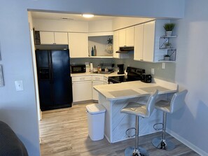A modern kitchen with white cabinets, a black refrigerator, a microwave, a stove, and a sink. There are two bar stools and a trash can.