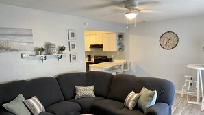 A modern living room with a dark grey sectional sofa, a round ottoman, and a dining area with a white table and chairs. A ceiling fan and a wall clock are visible. - Located right outside of the Vibe District at the Virginia Beach Oceanfront (Virginia Beach)