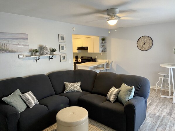 A modern living room with a dark grey sectional sofa, a round ottoman, and a dining area with a white table and chairs. A ceiling fan and a wall clock are visible.