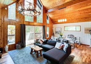 A spacious living room with a large sectional sofa, a wooden coffee table, and a dining area with a table and chairs. Large windows provide ample natural light and views of the outdoors. - Boho, Modern-Rustic A-Frame Cabin 5 Min to Village (Lake Arrowhead)