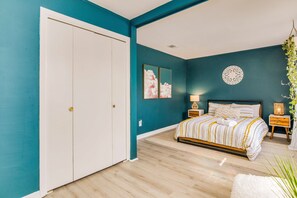 A bedroom with teal walls, a wooden floor, a white wardrobe, a bed with striped bedding, two bedside tables with lamps, and a decorative wall piece.