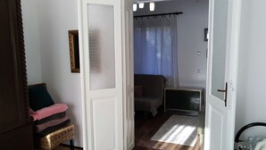 Une pièce avec un plancher en bois, une porte blanche dotée d'un panneau en verre dépoli, un banc en osier, un canapé marron, une petite table et une fenêtre avec des rideaux.