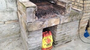 Un barbecue extérieur en briques, doté d’une grille en métal, un sac de charbon de bois et un balai appuyé contre le mur.