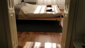 Une petite chambre avec un lit simple, un plancher en bois, un lit recouvert d’une couette blanche et une fenêtre ornée de rideaux légers.