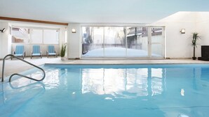 Una piscina interior con paredes de vidrio transparente, sillas azules y vistas a un área exterior nevada.