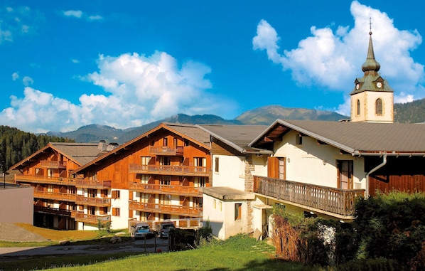 Un immeuble en bois à plusieurs étages, doté de balcons, une église avec un clocher et des montagnes en arrière-plan.