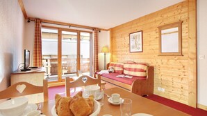Una habitación acogedora con detalles de madera, una mesa de comedor dispuesta con platos, tazas y un bol de pasteles. Hay un sofá con una manta de cuadros y una lámpara sobre una mesita.