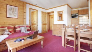 Una habitación acogedora con suelo de madera, un sofá, una mesa de comedor con sillas, una computadora portátil y una zona de cocina con armarios de madera.