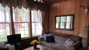 Indretning i en hyggelig hytte med en træsofa, et sofabord med en vase solsikker, et fjernsyn og et stort vindue med gardiner.