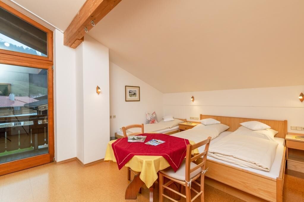 A room with a bed, a table with a red tablecloth, and a window with a view of a house.