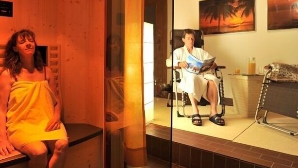 Een sauna met houten plafond, een glazen wand en een persoon die in een stoel een boek leest.