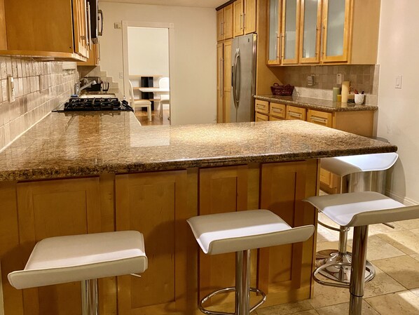 Eine Küche mit Granit-Arbeitsplatten, Holzschränken, einem Edelstahl-Kühlschrank und drei weißen Barhocker.