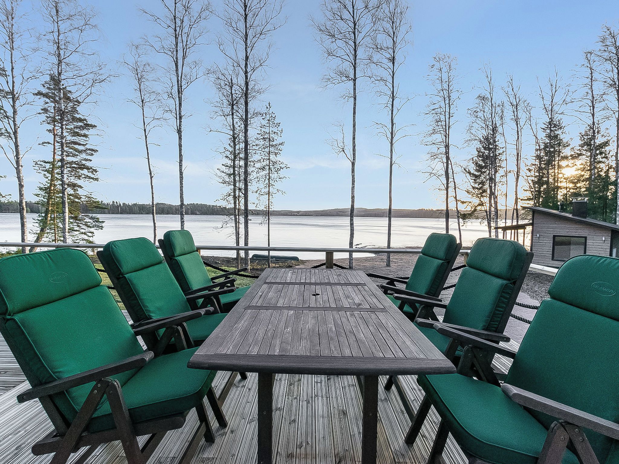 A deck with a wooden table and green chairs, overlooking a body of water and trees.