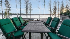A deck with a wooden table and green chairs, overlooking a lake with trees and a cabin in the background. - Metsola by Interhome (Ruovesi)