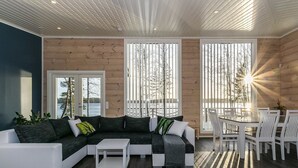A modern living room with a sectional sofa, a coffee table, and a dining area with white chairs. Large windows provide views of the outdoors. - Metsola by Interhome (Ruovesi)