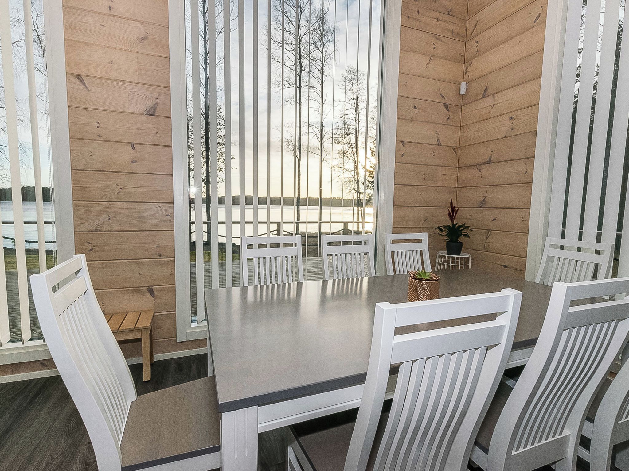 A wooden cabin interior with a dining table and white chairs, a small wooden bench, and a potted plant.